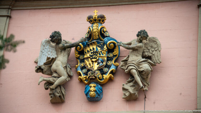 Crowned blue-and-gold coat of arms mounted on a pink wall and flanked by two sculpted winged figures.