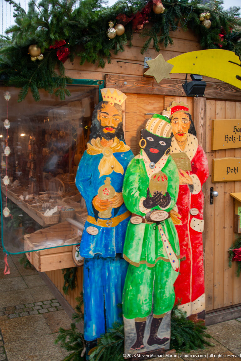 Three painted wooden figures in blue, green, and red traditional-style costumes beside a decorated Christmas market stall.