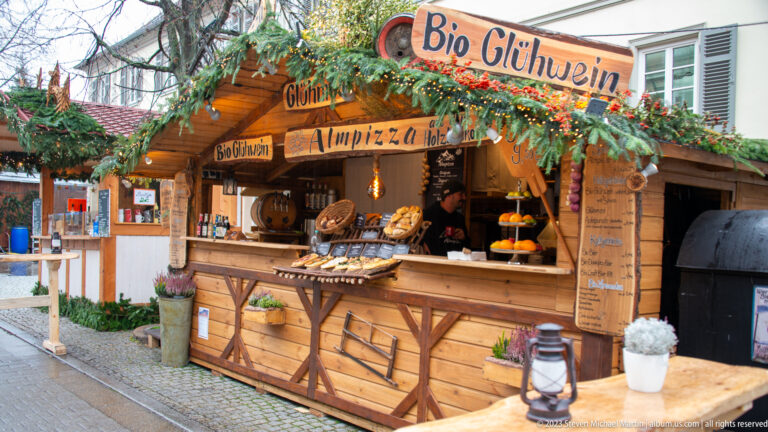 Wooden Christmas market stall labeled Bio Glühwein, decorated with evergreen branches, menus, hanging pretzels, and a service counter.