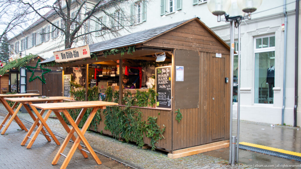 Small wooden Christmas market food stall with evergreen trim, menu boards, warm interior lights, and standing tables outside.