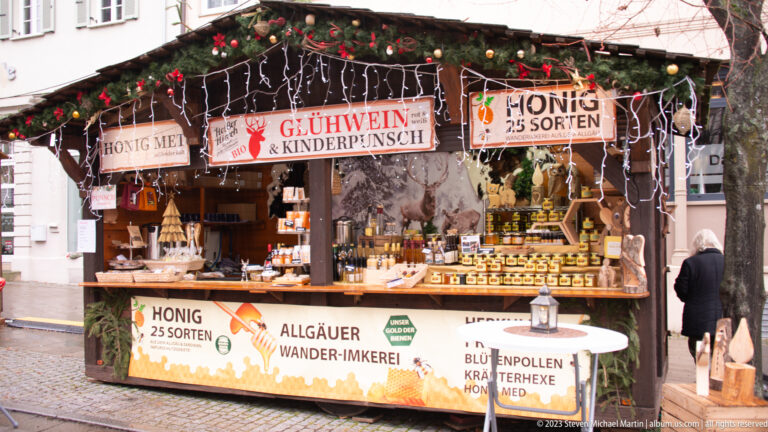 Christmas market stall displaying rows of honey jars with signs for 25 varieties, Allgäu beekeeping, Glühwein, and Kinderpunsch.