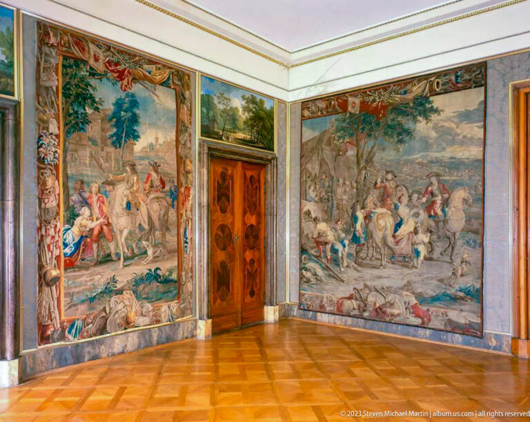 Corner of a palace room with two large narrative tapestries, inlaid wooden door, pale trim, and parquet floor.
