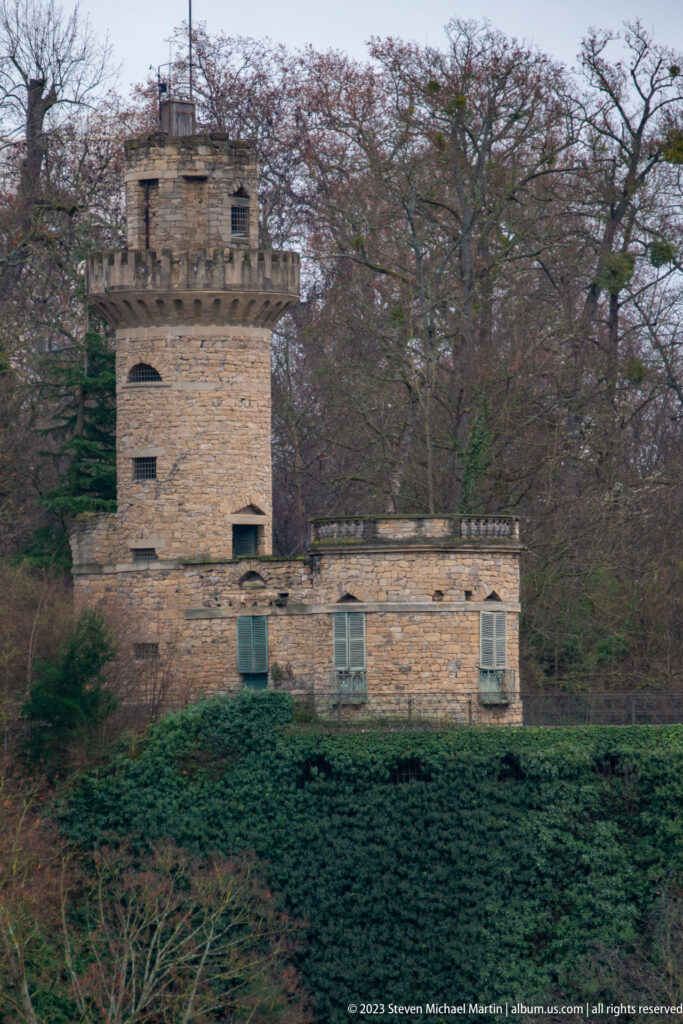 Small stone tower attached to a low masonry building among trees and hedges in Ludwigsburg parkland.