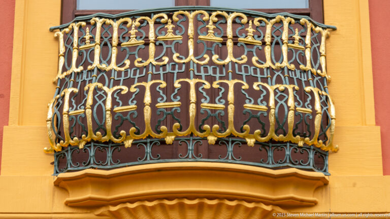 Close view of a curved balcony with dark ironwork and gilded scrolls on an ocher-and-red palace façade.