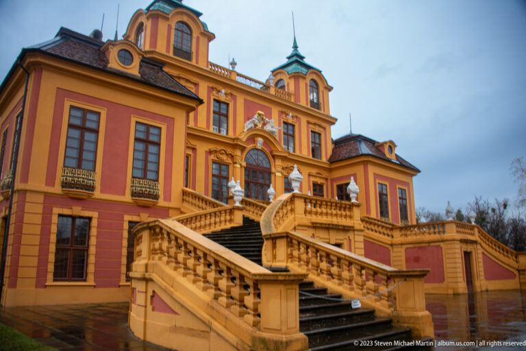 Angled view of Schloss Favorite with broad exterior staircase, balustrades, sculptural finials, and red-and-ocher façade.
