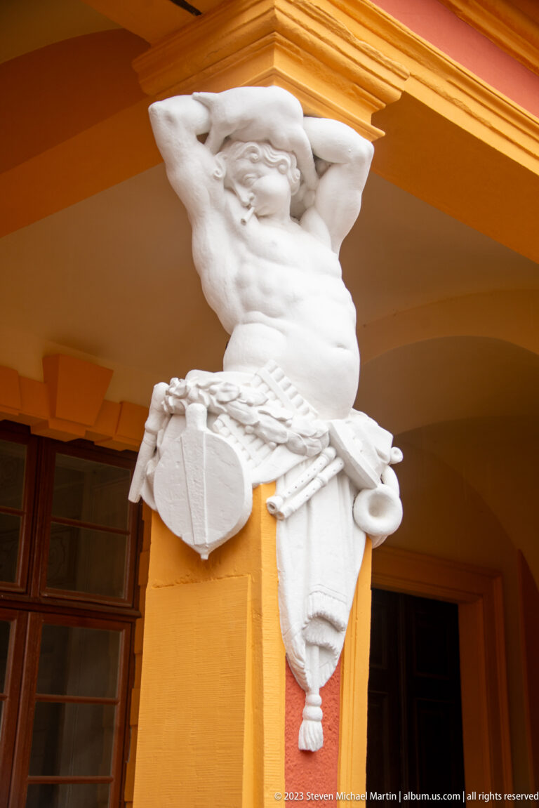 White male half-figure with shield and military-style attributes projecting from an ocher arcade pier at Schloss Favorite.
