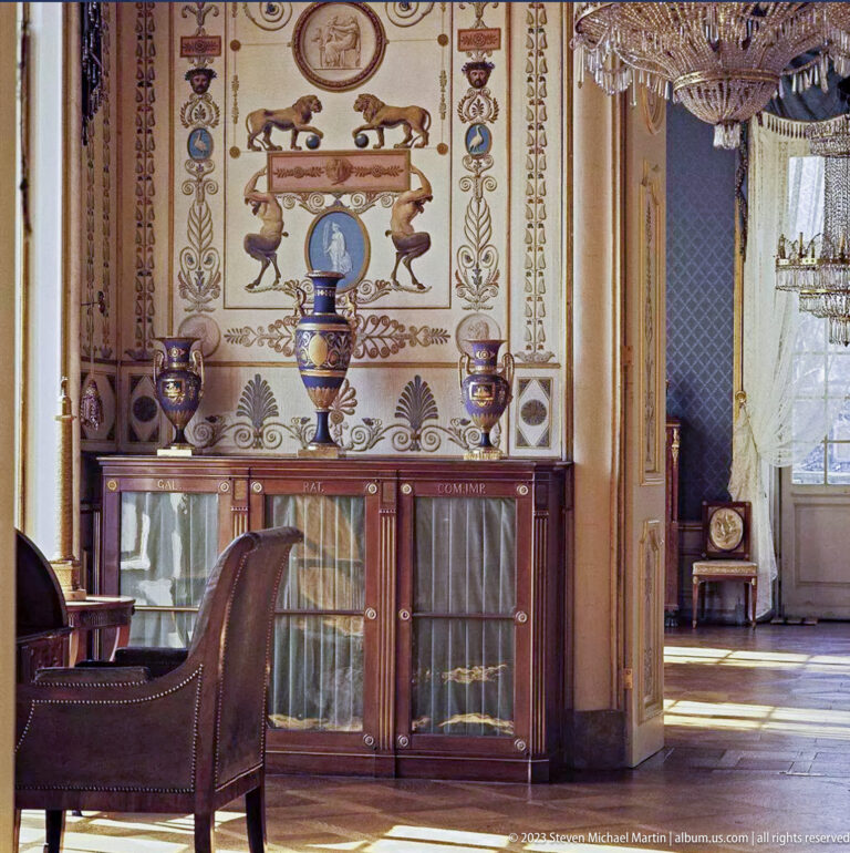 Decorated palace room with painted wall ornament, dark glazed cabinet, ceramics, chair, and open doorway to another chamber.