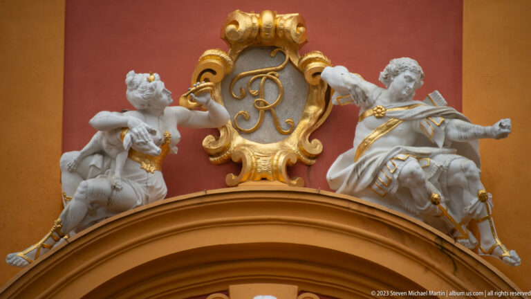 Two white sculpted figures flank a gilded monogram cartouche above an arched opening at Schloss Favorite.