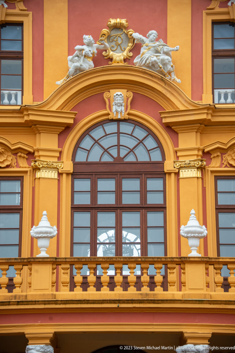 Large arched window, balcony balustrade, pilasters, gilded ornament, and sculptural crest on Schloss Favorite.