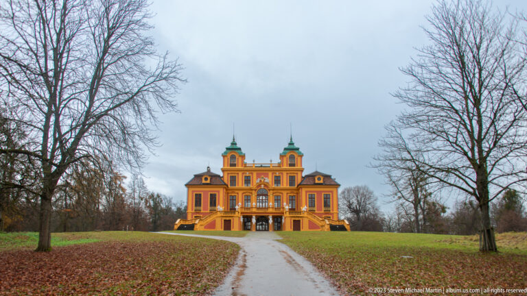 Distant Schloss Favorite centered at the end of a park road between leafless trees and winter lawns.