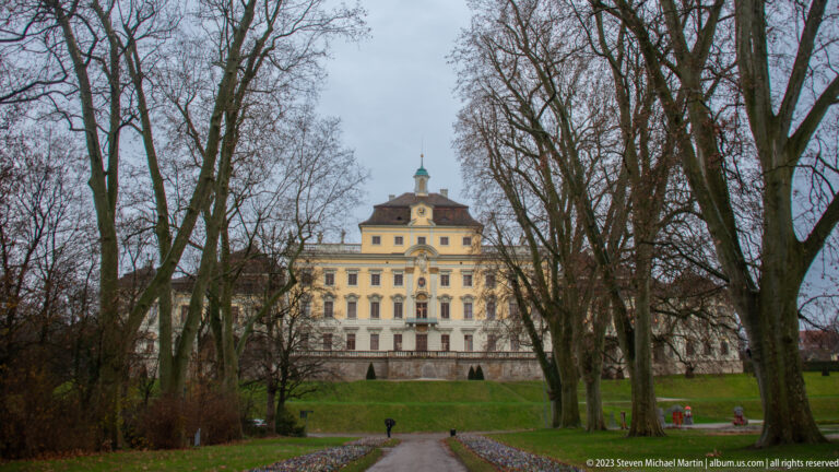 Pale symmetrical Baroque palace with central pavilion and roof cupola seen through leafless trees at the end of a path.