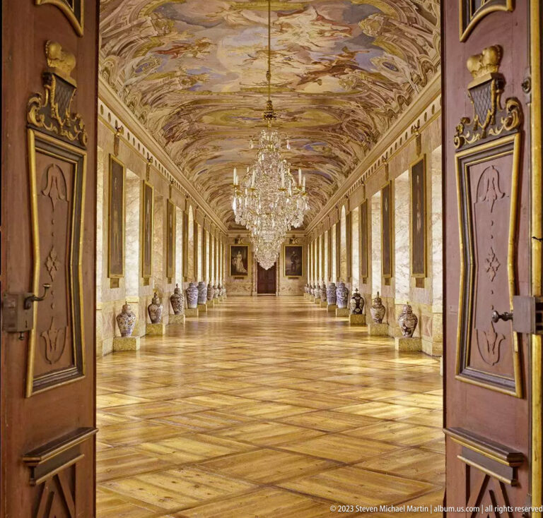 Long palace gallery with painted ceiling, chandeliers, tall windows, wall panels, decorative vessels, and parquet floor.
