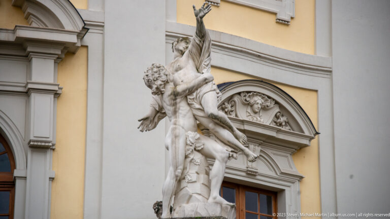 White sculptural group of intertwined figures beside an arched doorway on a pale-yellow palace façade.
