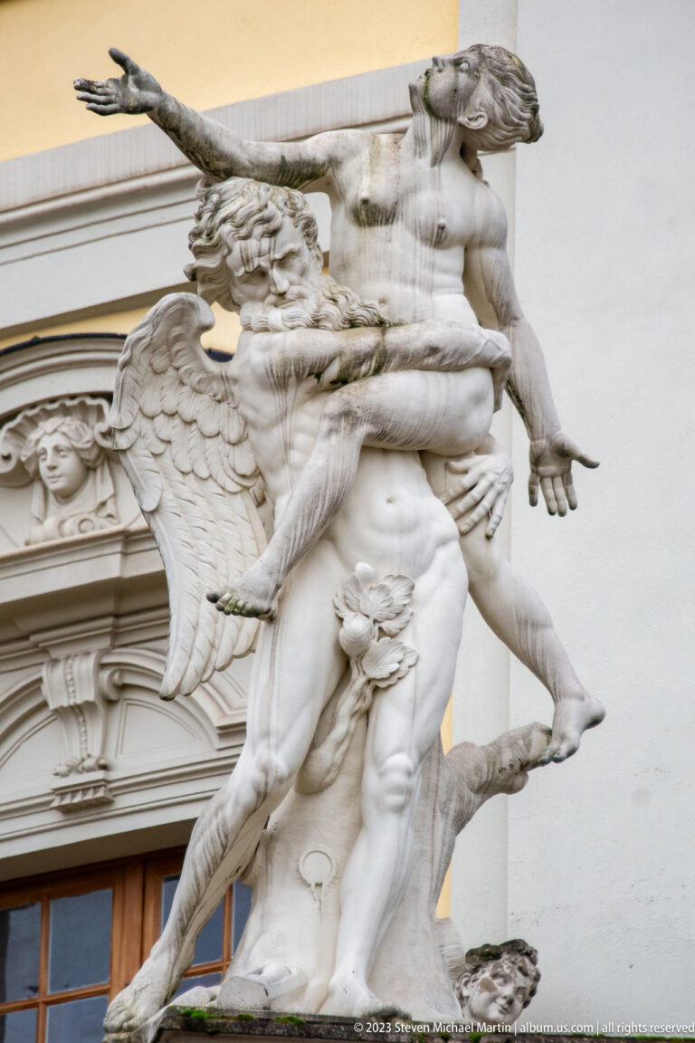 Close view of pale Baroque sculpture with overlapping figures, drapery, and a winged figure above a palace opening.