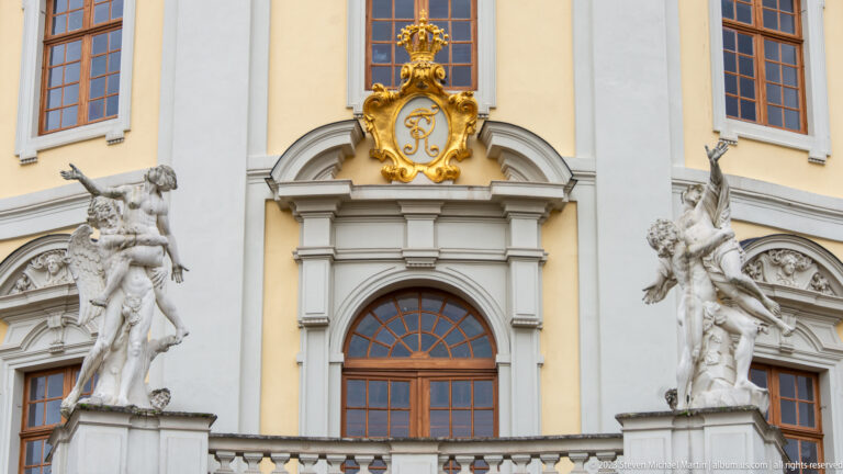 Arched palace window beneath a gilded monogram, flanked by large white sculptural groups on a pale-yellow façade.