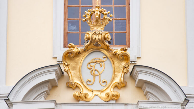 Gilded crowned monogram in an ornate cartouche above an arched opening on a pale yellow palace façade.