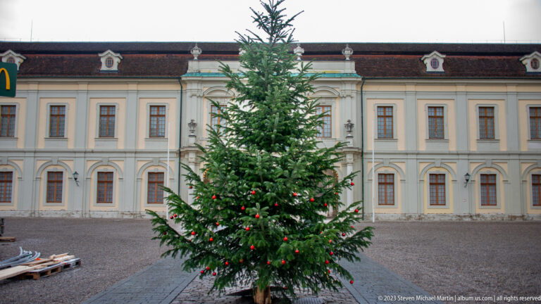 Decorated evergreen Christmas tree centered before a long pale palace wing with red-tiled roof and rows of windows.