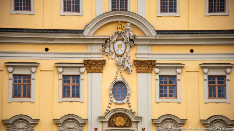 Crowned heraldic cartouche and cascading sculptural ornament between windows on a yellow palace façade.