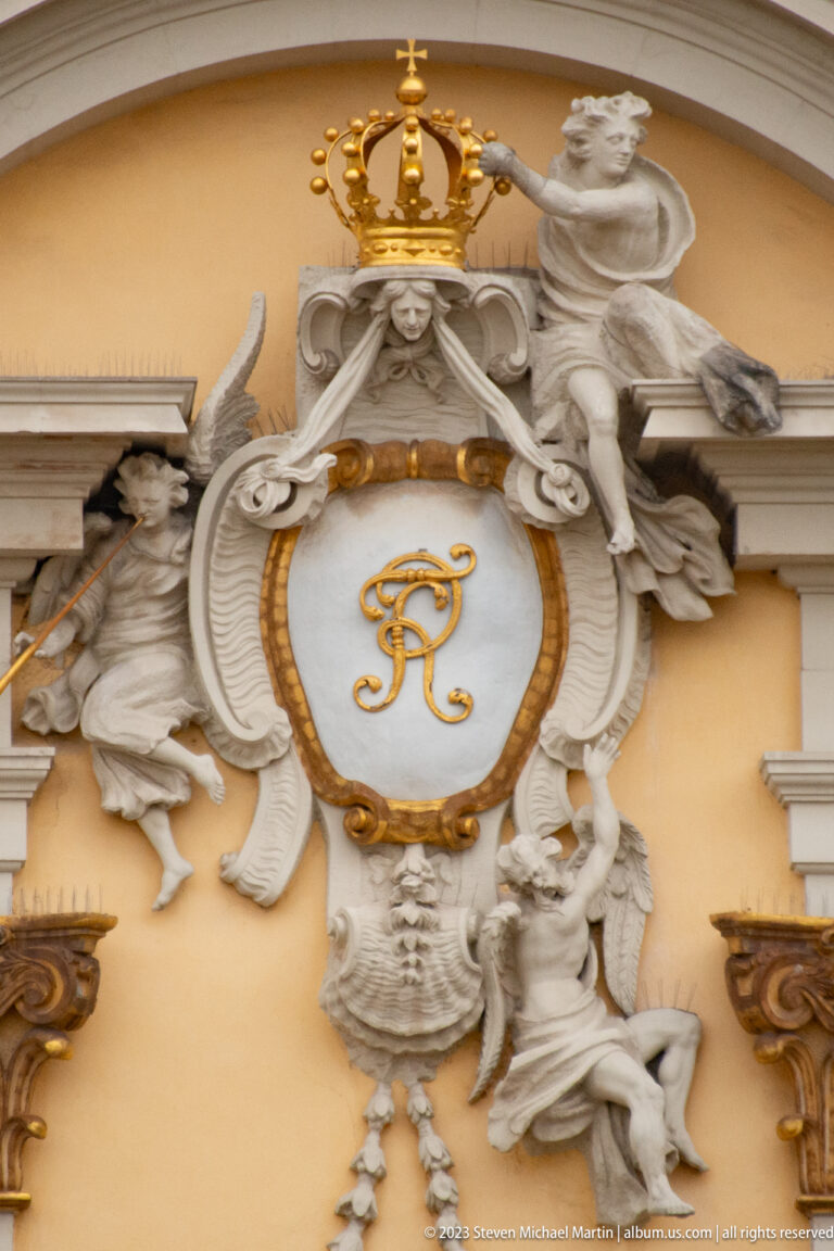 Large crowned gilded monogram in an oval shield surrounded by sculpted figures and scrolling Baroque ornament.