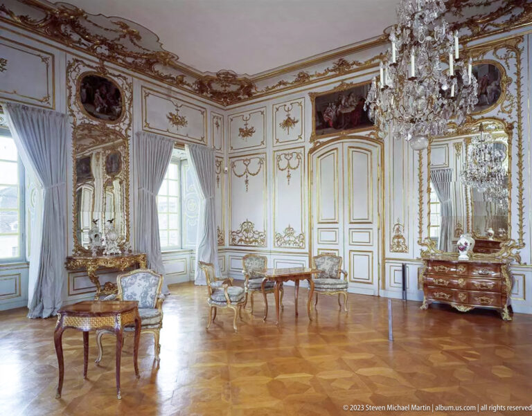 White-and-gold palace salon with crystal chandelier, mirrors, carved paneling, painted overdoors, seating, and parquet floor.