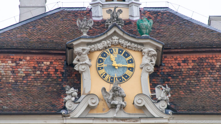 Blue-and-gold Roman-numeral clock set in a sculpted Baroque surround against a red-tiled roof.
