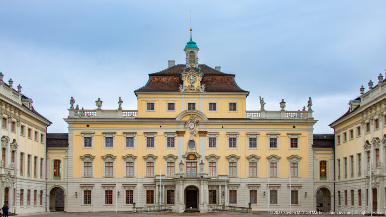 Symmetrical yellow palace façade with central clock, green cupola, rooftop statues, and formal doorway across a courtyard.