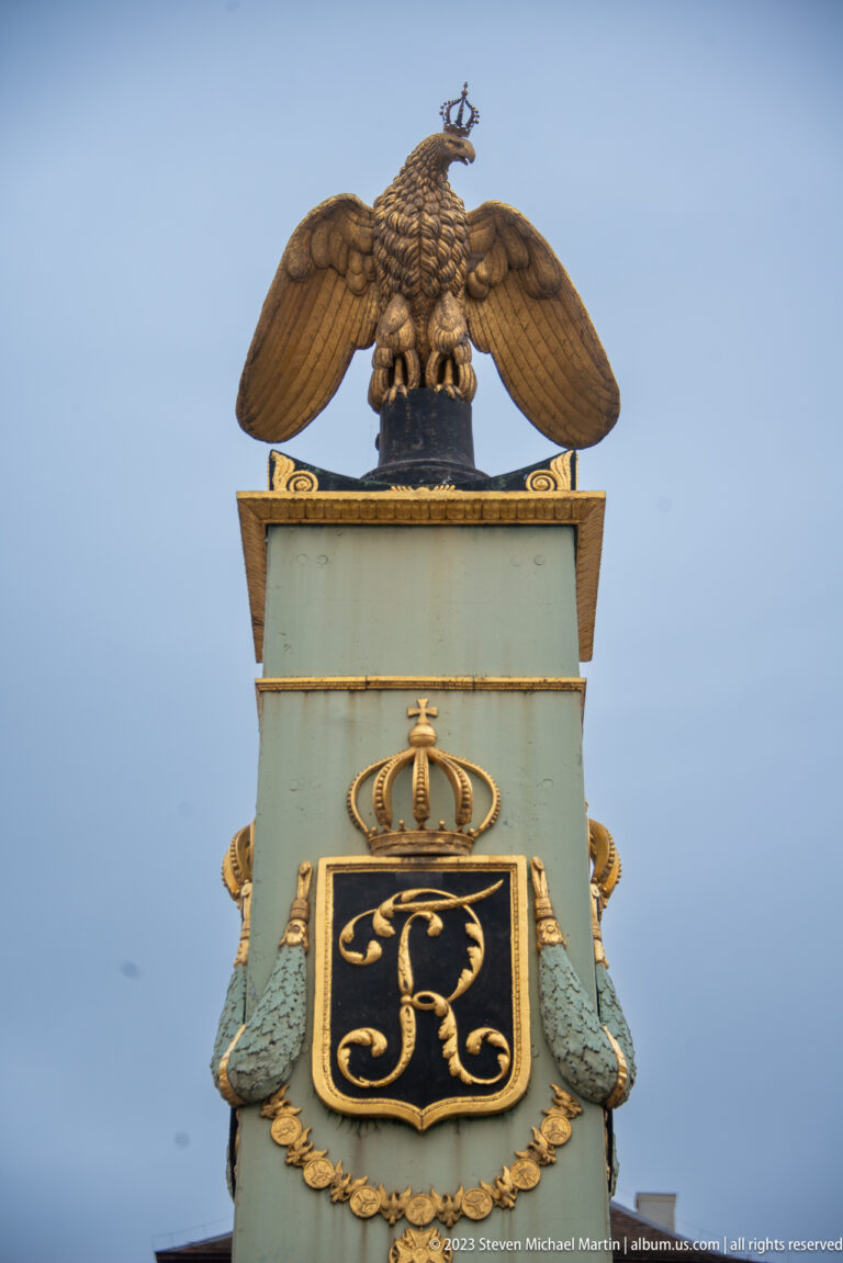 Winged eagle atop a pale green heraldic pedestal with black-and-gold crowned monogram panel.