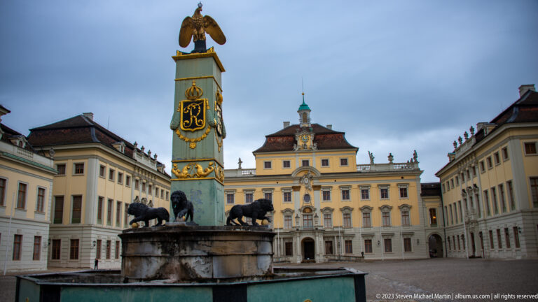 Tall green-and-gold eagle-topped courtyard monument aligned with the yellow palace façade behind it.
