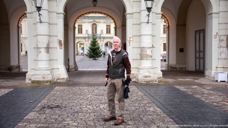 Man standing in a stone palace arcade with cobbled paving and a Christmas tree visible through the arch behind.