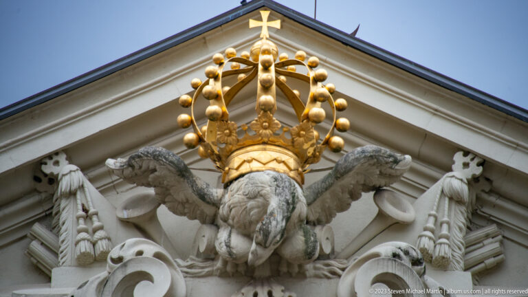 Gilded crown above a sculpted eagle and carved trophies in the triangular pediment of a palace building.