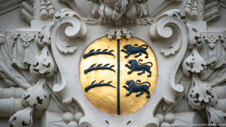 Gold oval coat of arms with black antlers and lions surrounded by elaborate pale stone scrollwork.