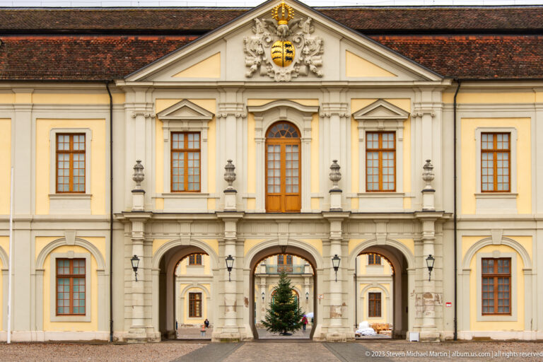 Three-arched palace gateway with upper windows, heraldic pediment, and a Christmas tree visible through the center arch.