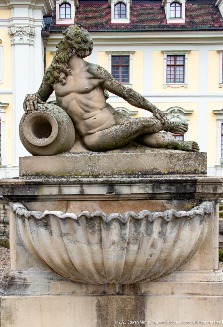 Weathered reclining male fountain figure beside a large vessel above a shell-shaped basin, with palace windows behind.