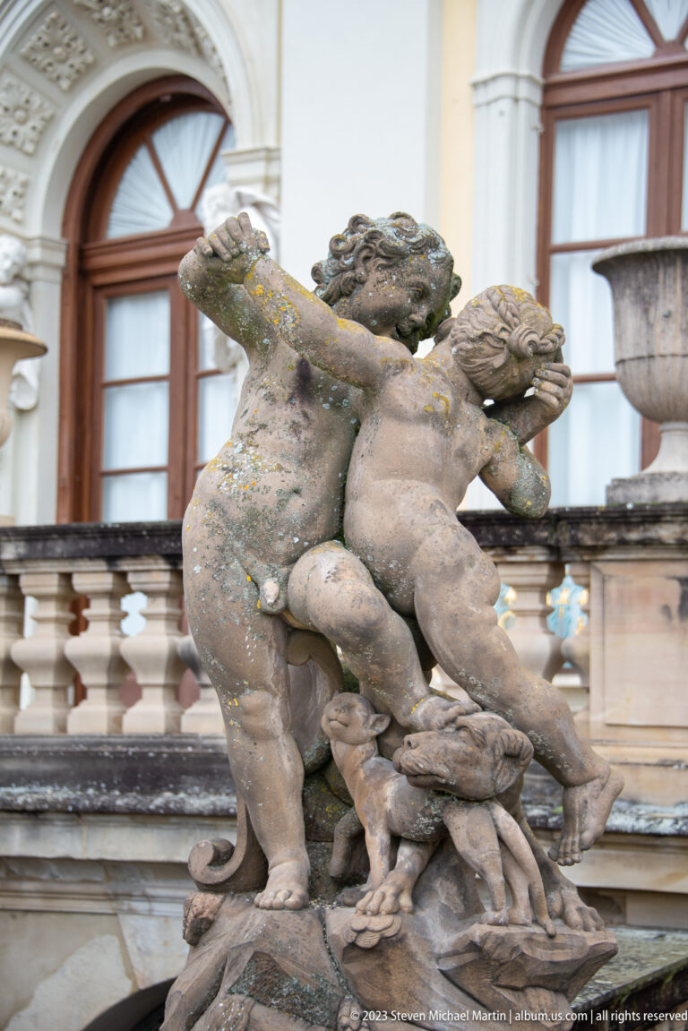 Two weathered stone putti twisting together in a palace fountain group, with windows and balustrade behind.