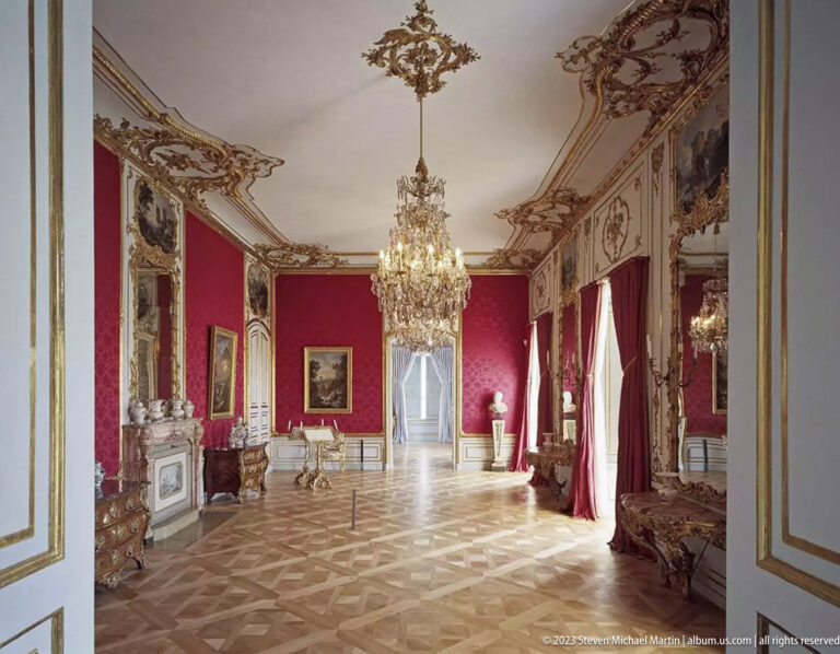 Formal red palace salon with chandelier, mirrors, paintings, historic furniture, tall windows, and parquet floor.