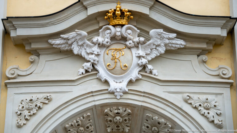 Pair of white sculpted eagles holding a crowned gilded monogram above an ornate arched doorway.