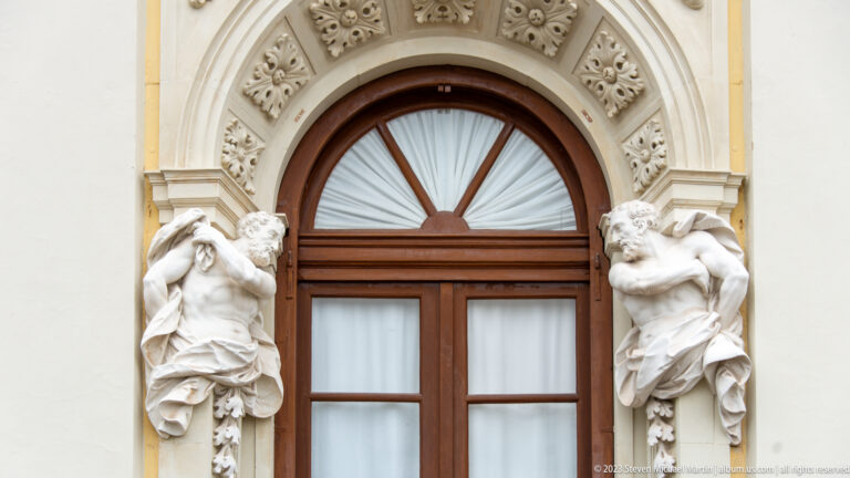 Arched wooden palace window flanked by two carved female figures and floral relief ornament.