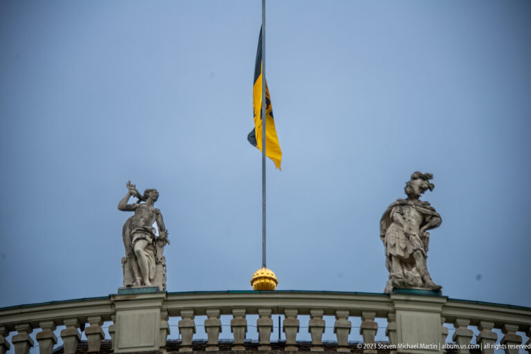 Two rooftop stone statues flanking a black-and-yellow flag on a palace balustrade under a gray sky.