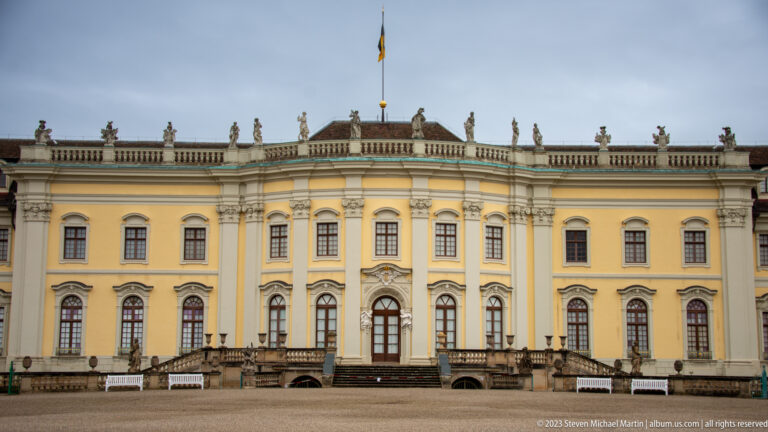 Curved yellow palace garden façade with tall windows, rooftop statues, central flagpole, and broad exterior stairs.