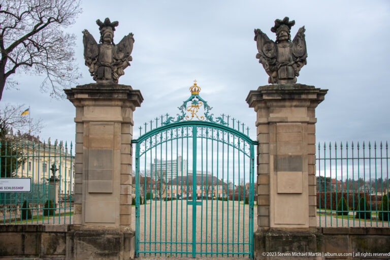 Turquoise wrought-iron palace gate between stone piers topped with sculptural trophies, with formal grounds beyond.