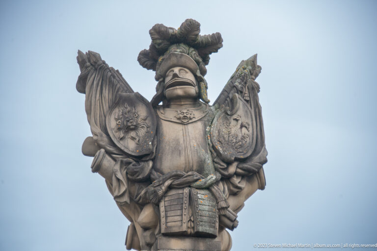 Weathered armored stone figure with shields and trophy forms crowning a palace gate pier against the sky.