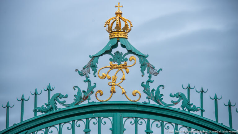 Gilded crown and monogram above turquoise wrought-iron gate scrollwork and spear-point finials.