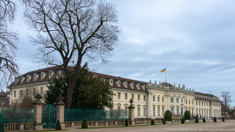 Long yellow palace wing seen obliquely behind a green fence and winter trees, with repeated dormers and windows.