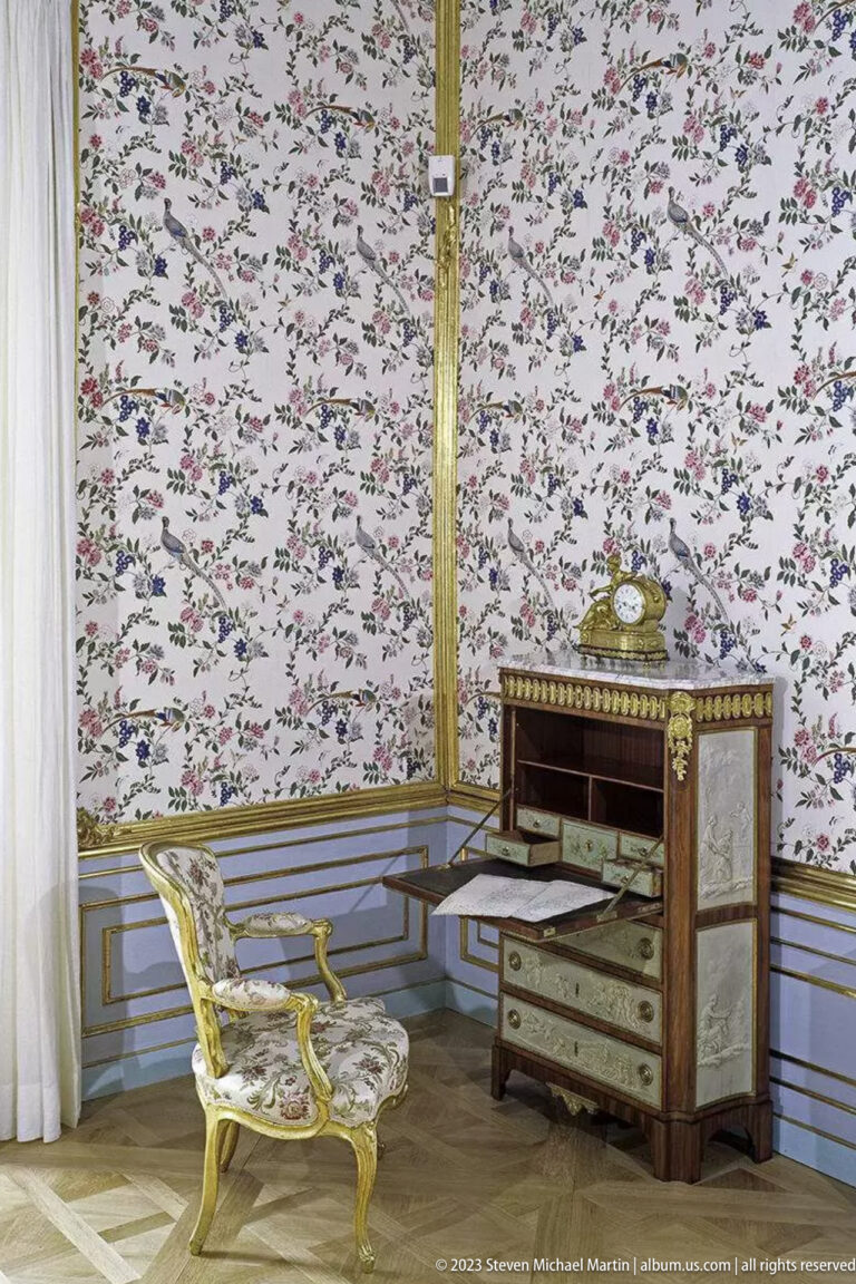 Floral-papered palace room with gilded armchair, dark commode with pale panels, and gold-trimmed wall molding.