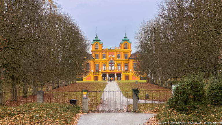 Bright yellow-orange Schloss Favorite with twin corner towers at the end of a straight path lined by leafless trees.