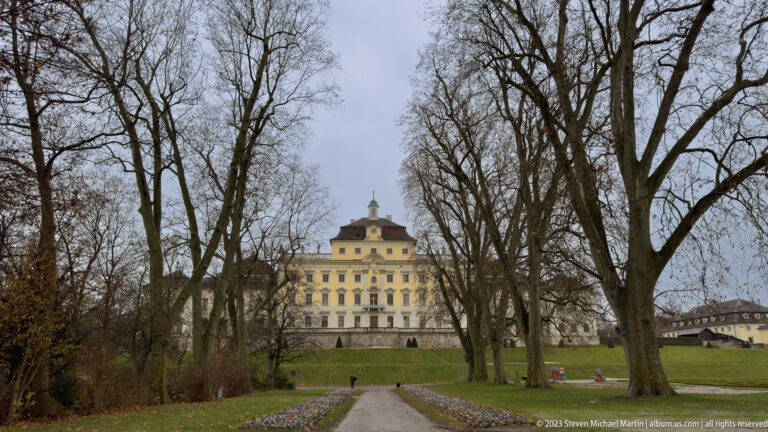 Distant pale palace building seen through a winter park of leafless trees with a straight path leading toward it.