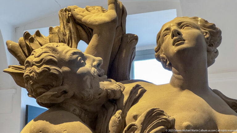Close view of a pale sculptural group with an upward-looking female figure beside a grotesque or satyr-like companion.