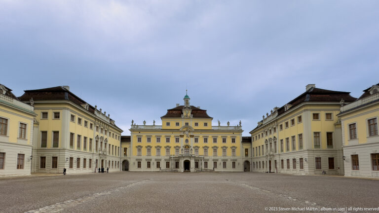 Wide gravel courtyard enclosed by pale yellow palace wings and an ornate central pavilion under an overcast sky.