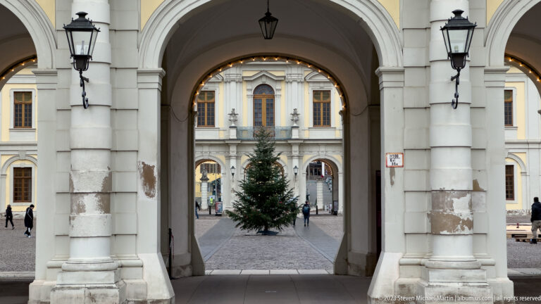 Rounded palace archway with black lanterns framing a Christmas tree and yellow façade in the courtyard beyond.