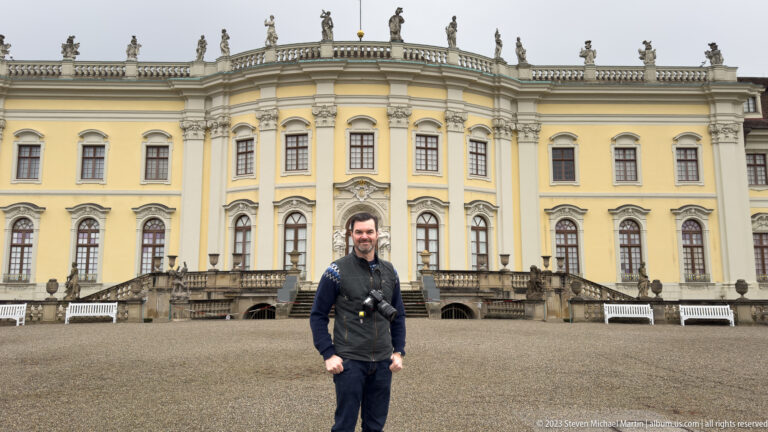 Visitor standing on a gravel court before an ornate yellow palace façade with tall windows, pilasters, and roof statues.